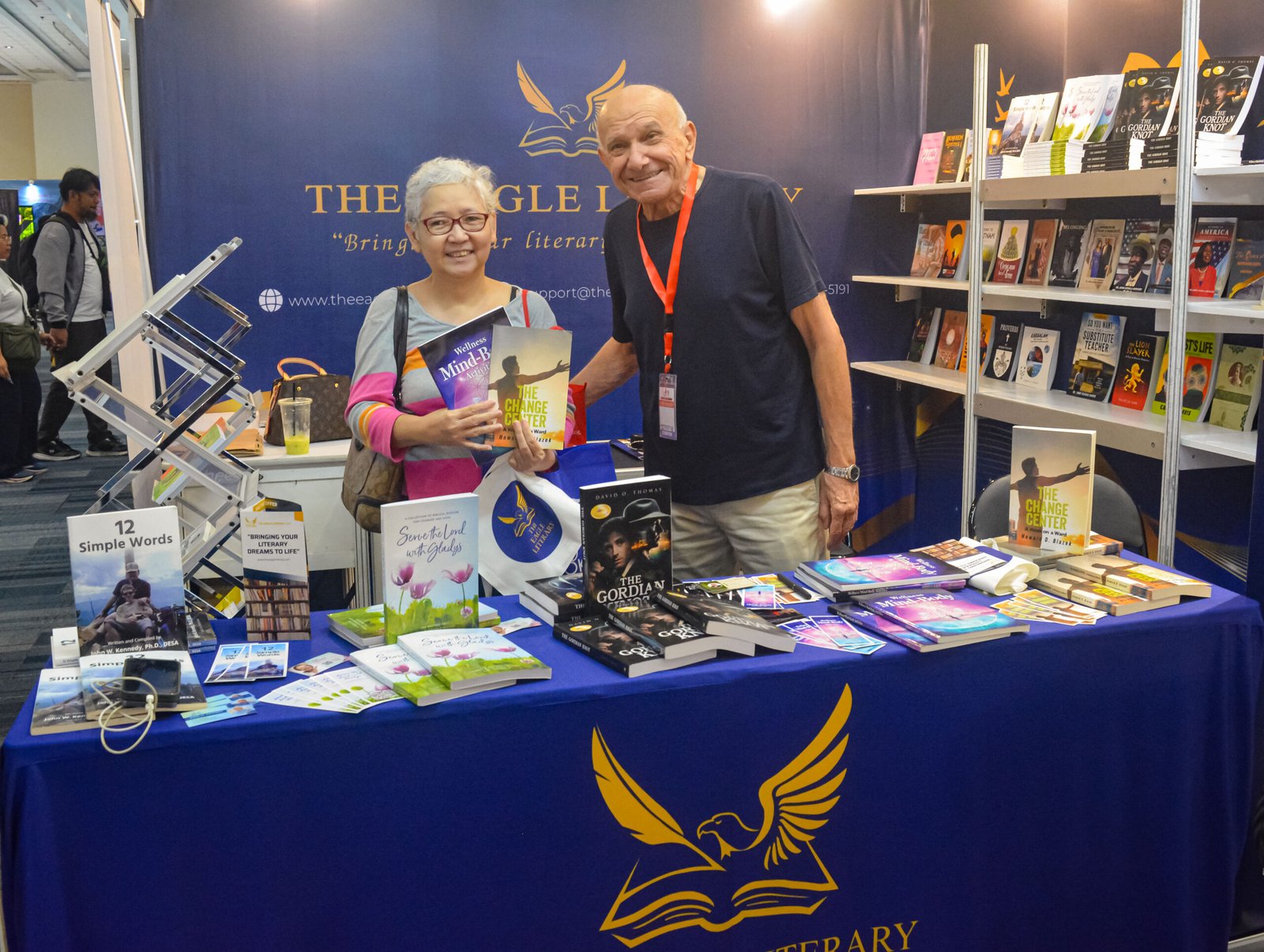 Two visitors holding books and smiling at The Eagle Literary booth during the 2026 Manila International Book Fair, surrounded by displayed books and promotional materials.