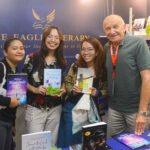 Four smiling attendees posing with books at The Eagle Literary's booth during the 2026 Manila International Book Fair, featuring book displays and promotional materials.