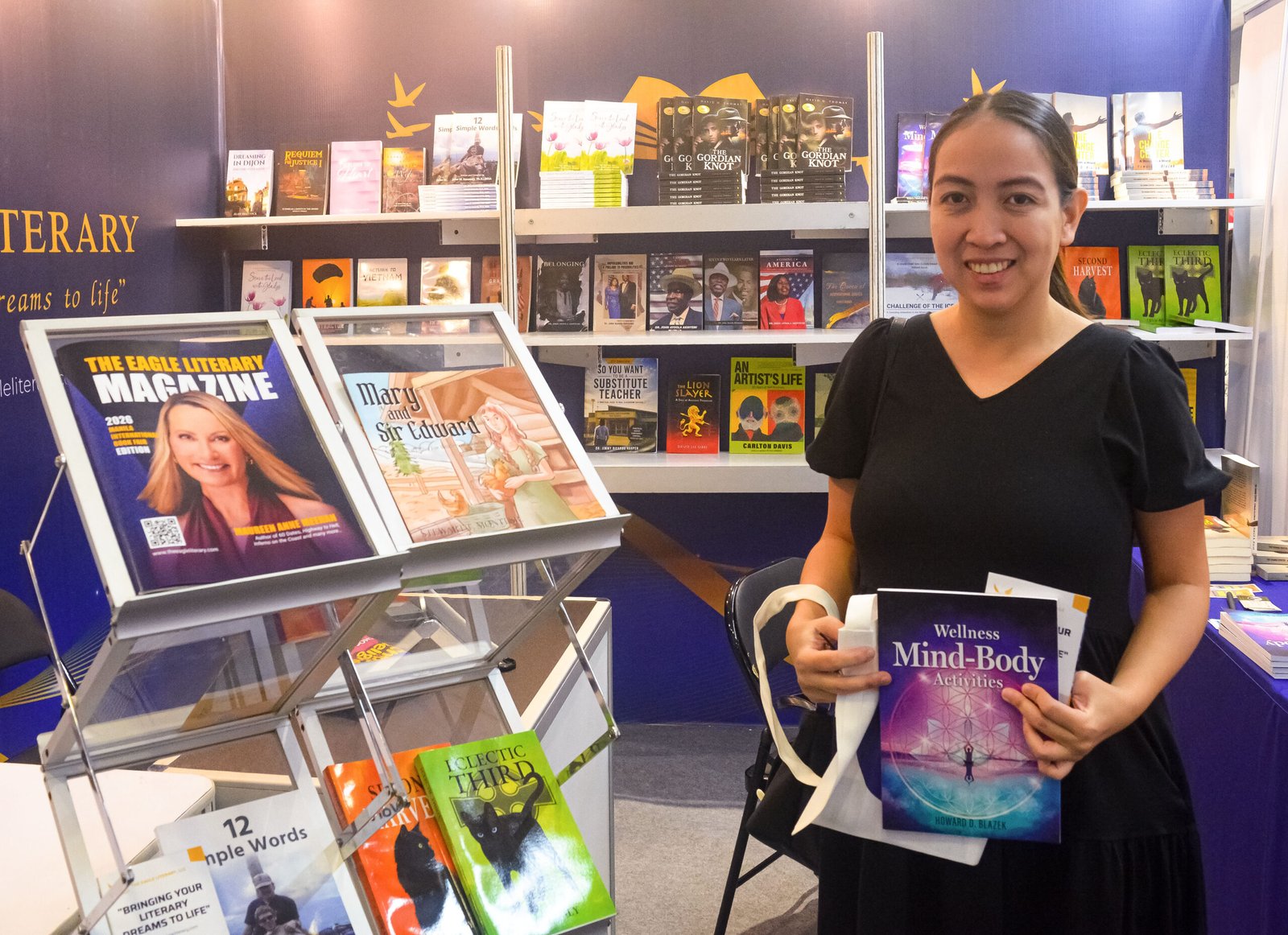 Smiling visitor holding the book Wellness Mind-Body Activities at The Eagle Literary's booth during the 2026 Manila International Book Fair, surrounded by book displays and literary magazines.