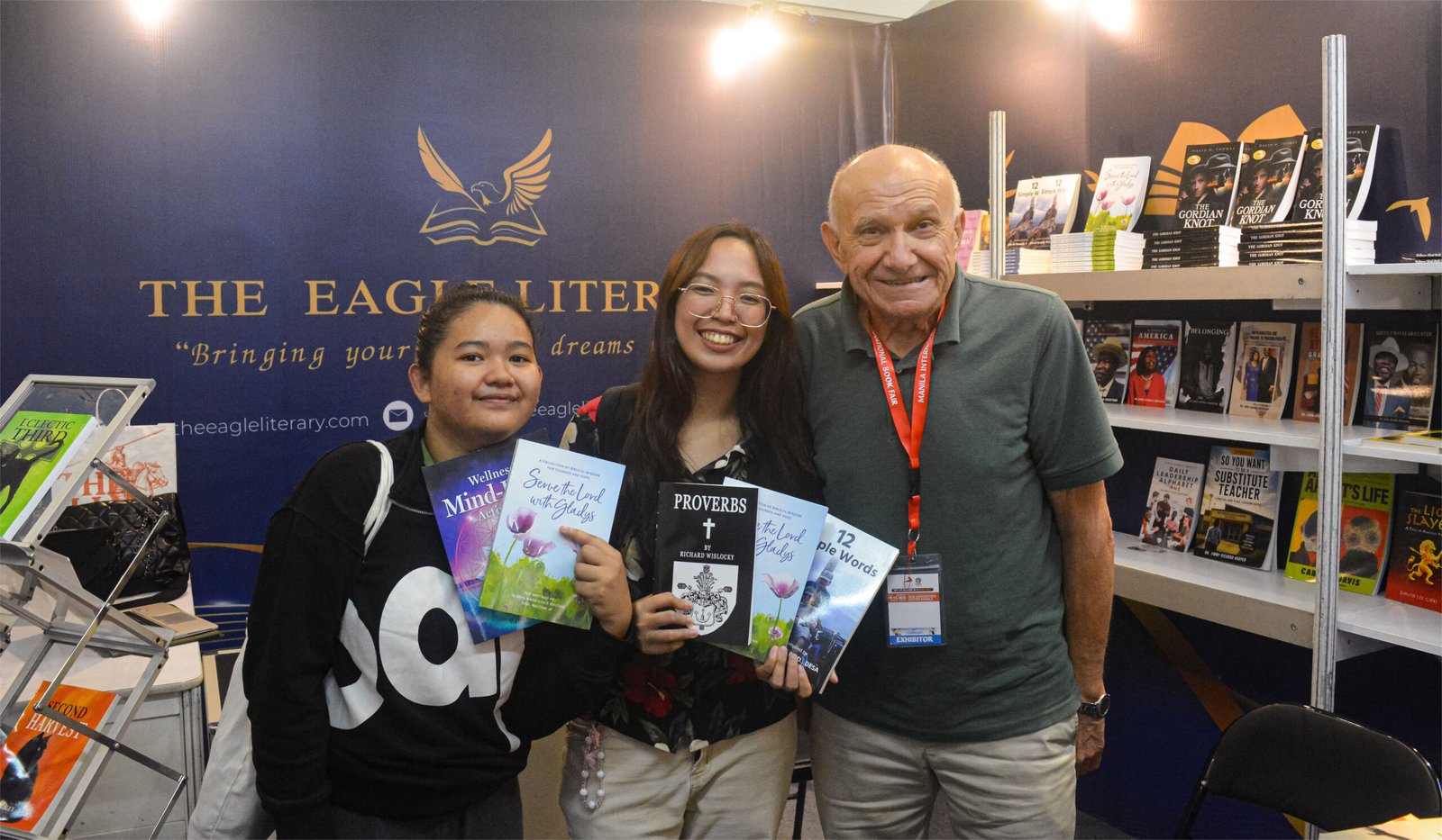 Three smiling attendees holding featured books at The Eagle Literary's exhibition booth during the 2026 Manila International Book Fair, surrounded by published titles and promotional displays.