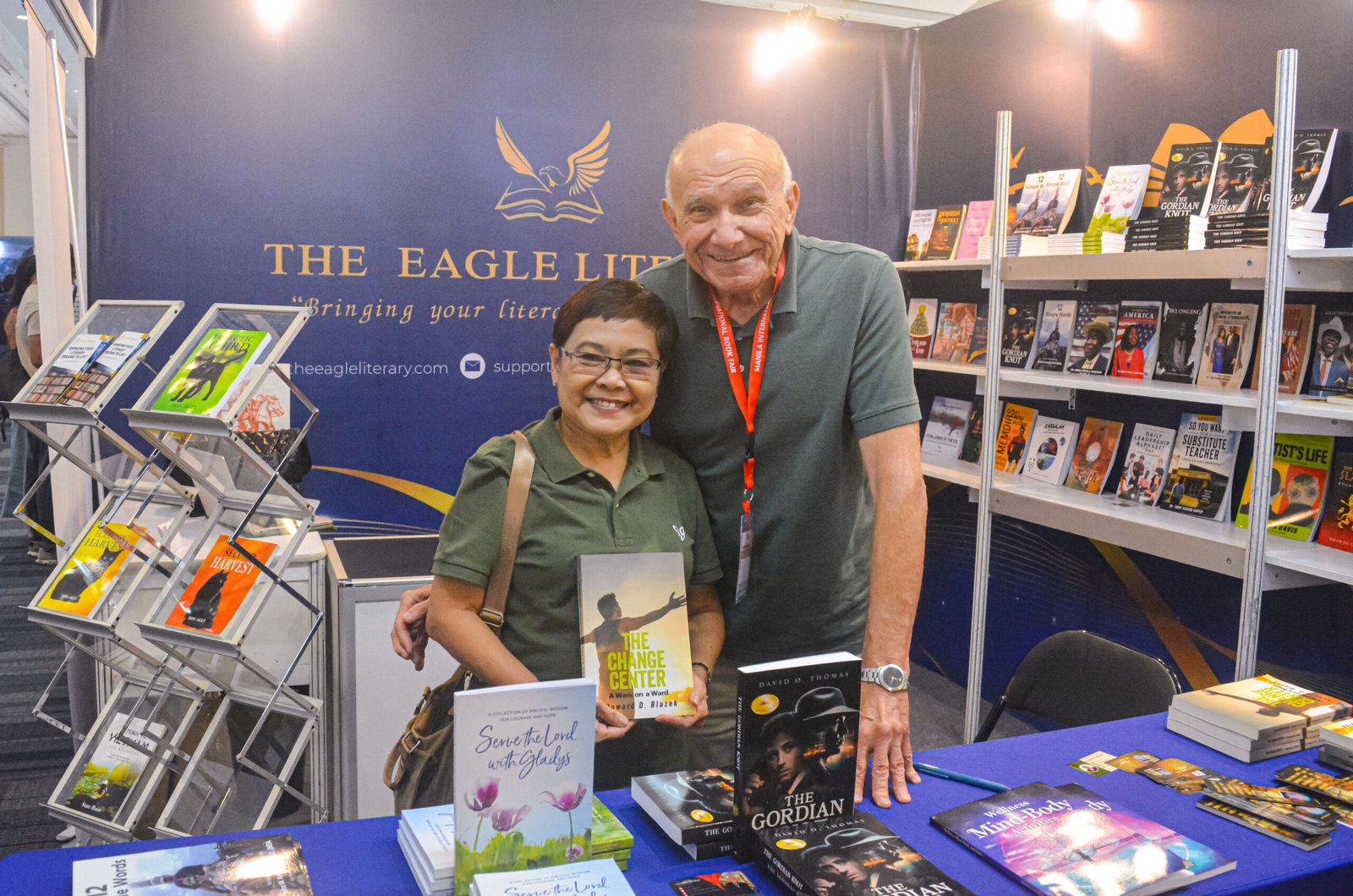 Two smiling attendees posing with a copy of The Change Center at The Eagle Literary's booth during the 2026 Manila International Book Fair, surrounded by featured books and literary displays.