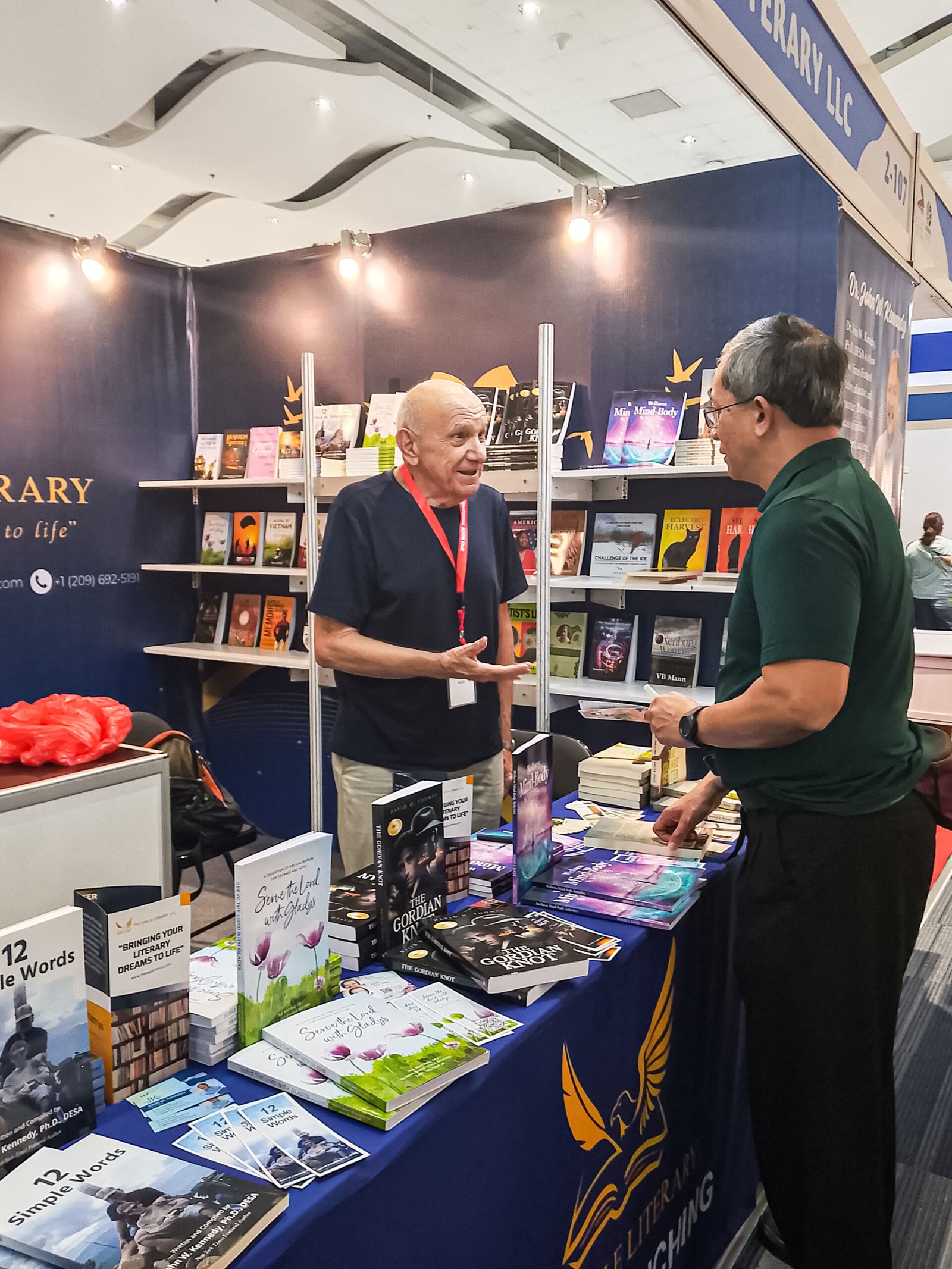 Two attendees engaging in a friendly conversation at The Eagle Literary's exhibition booth during the 2026 Manila International Book Fair, surrounded by featured books, promotional materials, and literary displays.