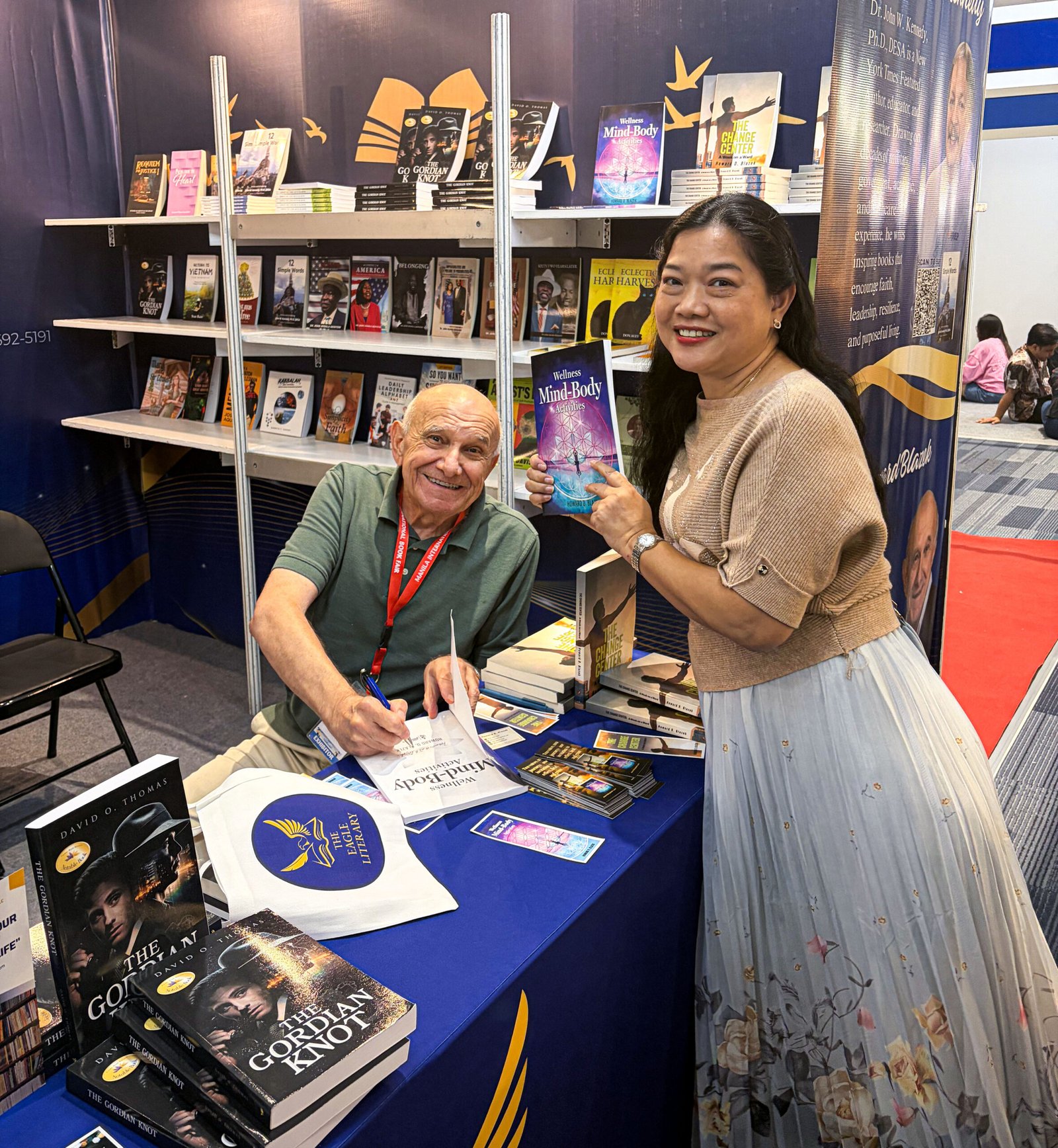 Smiling visitor holding Wellness Mind-Body Activities while an exhibitor signs a book at The Eagle Literary's booth during the 2026 Manila International Book Fair, surrounded by featured books and promotional displays.