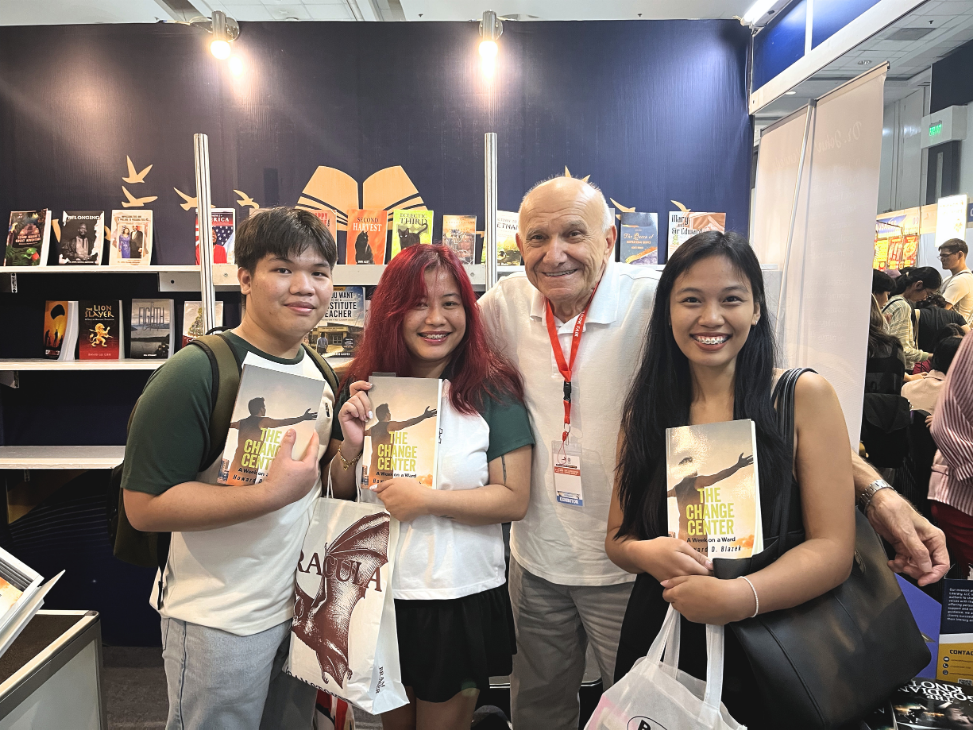 Four smiling attendees posing with copies of The Change Center: A Week on a Ward by Howard D. Blazek at The Eagle Literary's exhibition booth during the 2026 Manila International Book Fair, surrounded by featured literary publications.