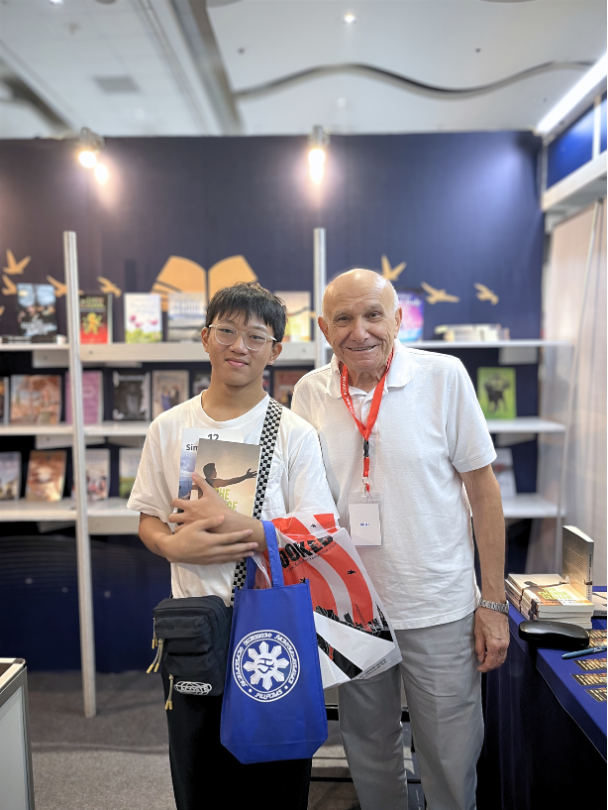 A young visitor holding a copy of 12 Simple Words poses with a smiling exhibitor at The Eagle Literary's booth during the 2026 Manila International Book Fair, surrounded by featured books and literary displays.